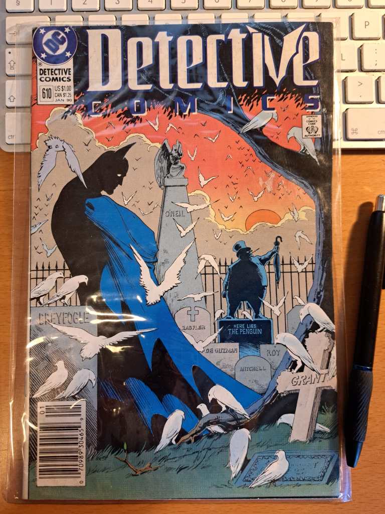 Photo of the Detective Comics 610, Jan 90 comic book, featuring Batman at a cemetery/graveyard, tombstones include Here Lies the Penguin, along others with thenames of Breyfogle, Raspler, De Guzman, Mitchell, Roy and Grant. Several pigeons in the scene. Computer keyboard and pen appear for scale. 